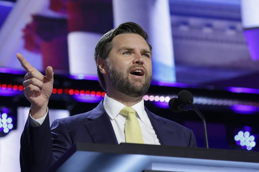 A bearded man dressed in a blue suit and yellow tie speaks at a lectern.