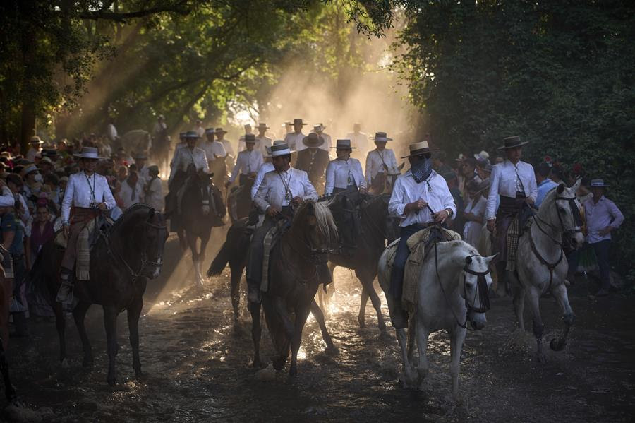 A group of people on horseback travel through a forest. The pilgrims on horseback are wearing white jackets and flat straw-woven hats. There are also people gathered on either side of the path watching the procession.