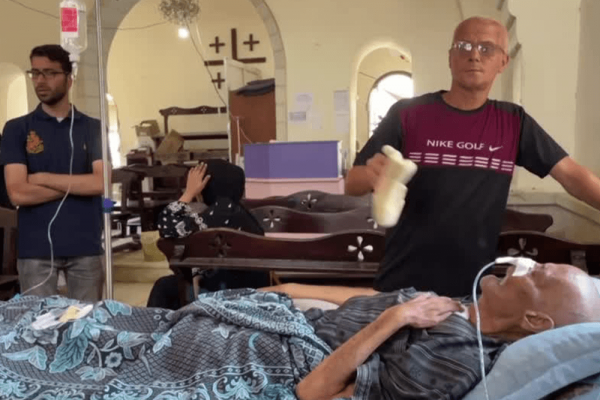 Older man lies in hospital bed connected to breathing tubes. Two other men stand watch nearby. 