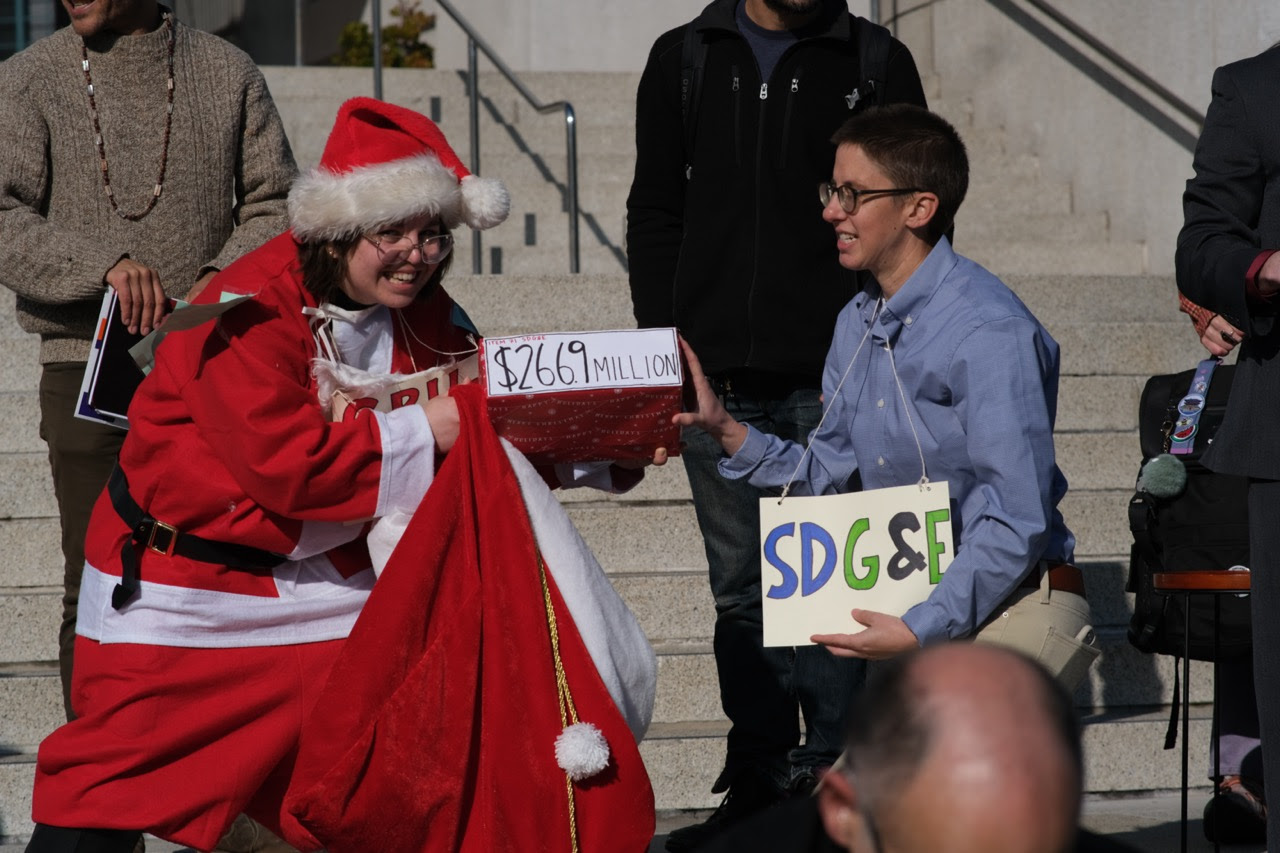 Picture of an attendee at rally against rate hike and decision to
keep Aliso Canyon Gas Facility open indefinitely wearing a CPUC Santa
costume. The attendee is smiling while handing over a box with a label
that says $266.9 Million on it to another rally attendee holding a
sign that says SDG&E.