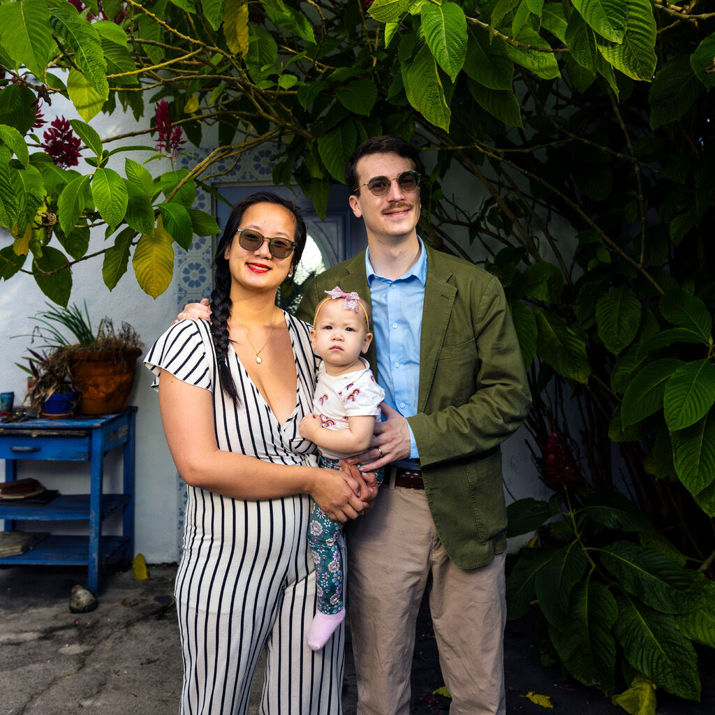 A couple holding a baby between them stand in a paved yard with greenery behind them.