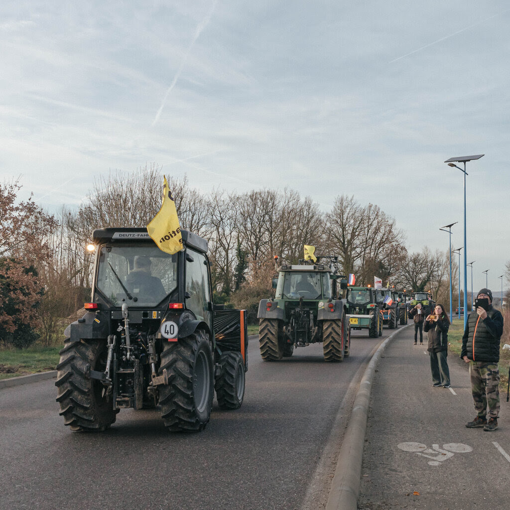 A line of tractors with yellow flags on a rural road. People beside the road are recording the procession via their cellphones.