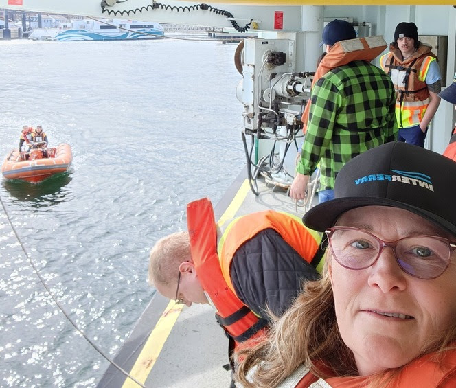 Person taking selfie with others on ferry wearing life vests and others in rescue boat in water in distance attached to the ferry by a rope