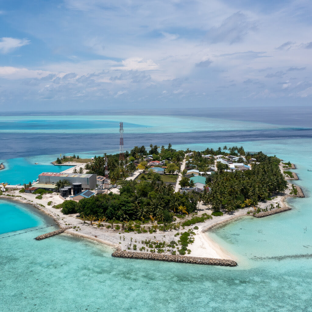 A wide shot of an island dotted with trees and a few buildings. The island is surrounded by turquoise blue waters.