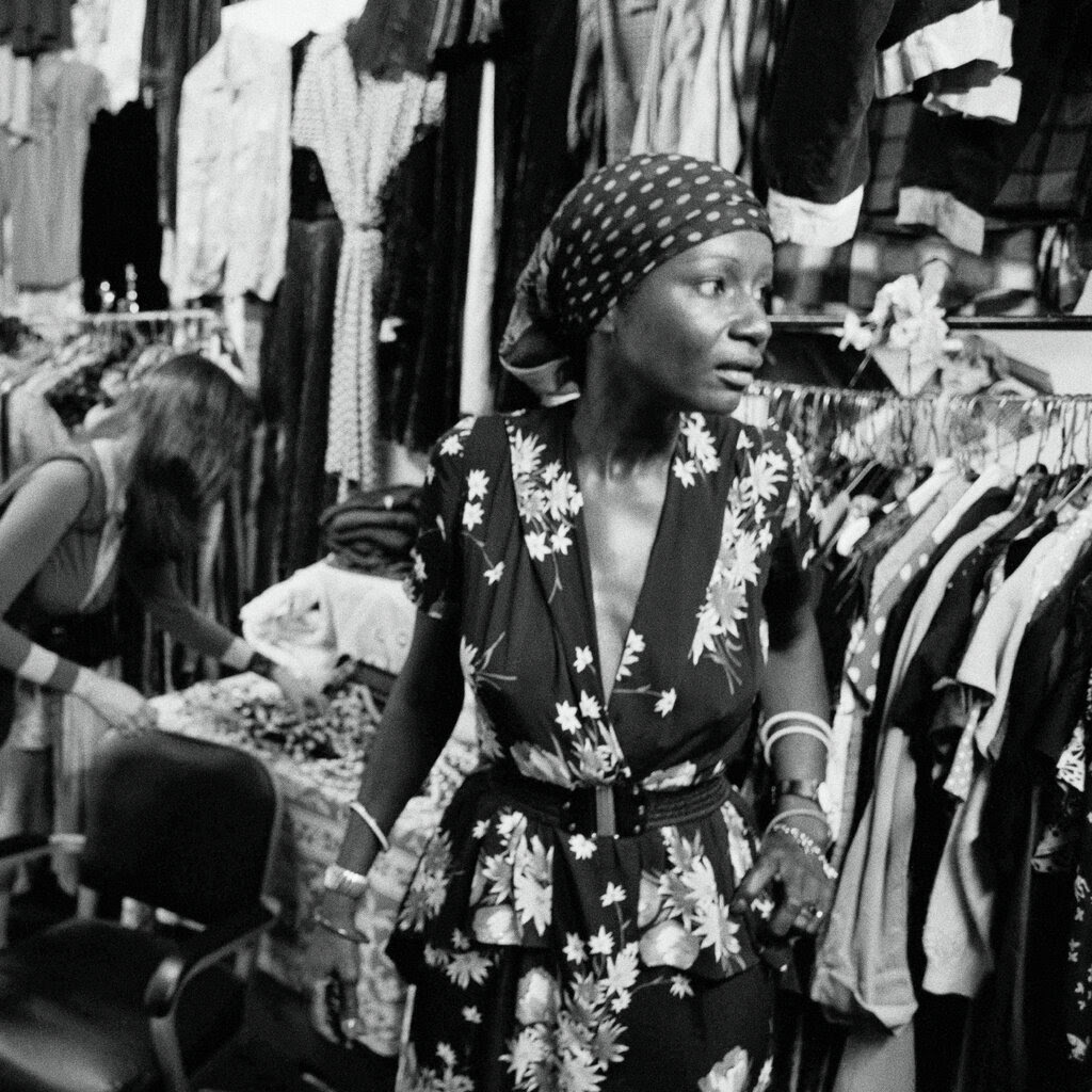 A black-and-white photo of a woman in a floral dress and a head wrap looking to her left in a clothing store while other people shop. 