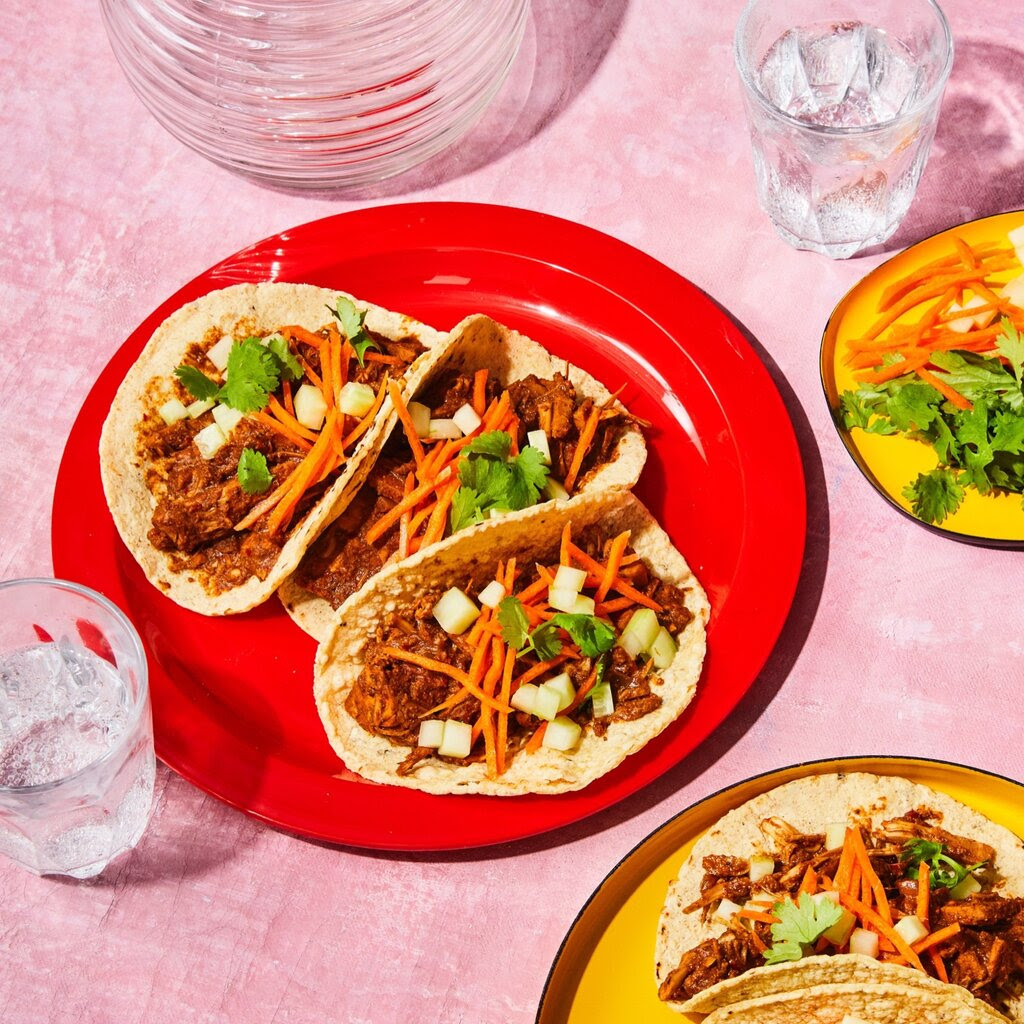 Jackfruit tacos on a red plate sit next to a small plate of toppings.