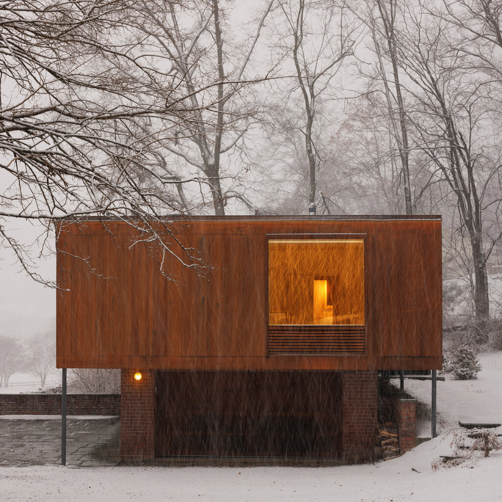 A side view of a boxy wooden house surrounded by bare trees and snow.