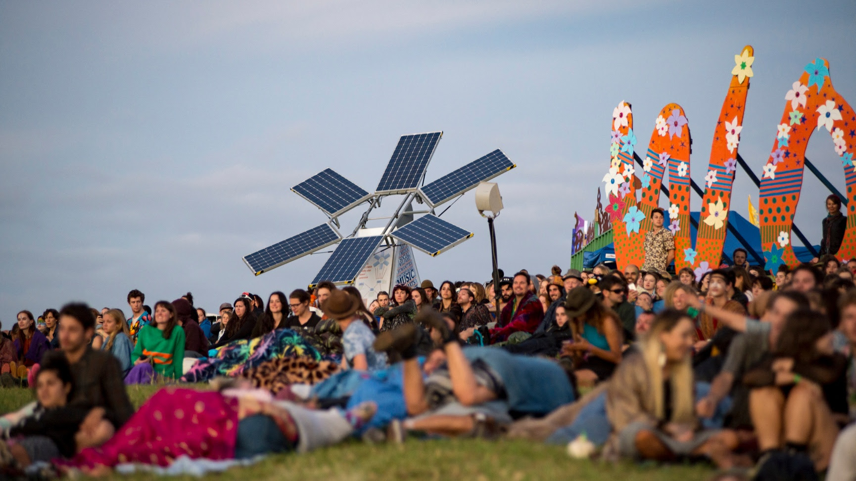 A solar panel structure in the shape of a flower, next to a large crowd of festival-goers sitting on a grass hill.