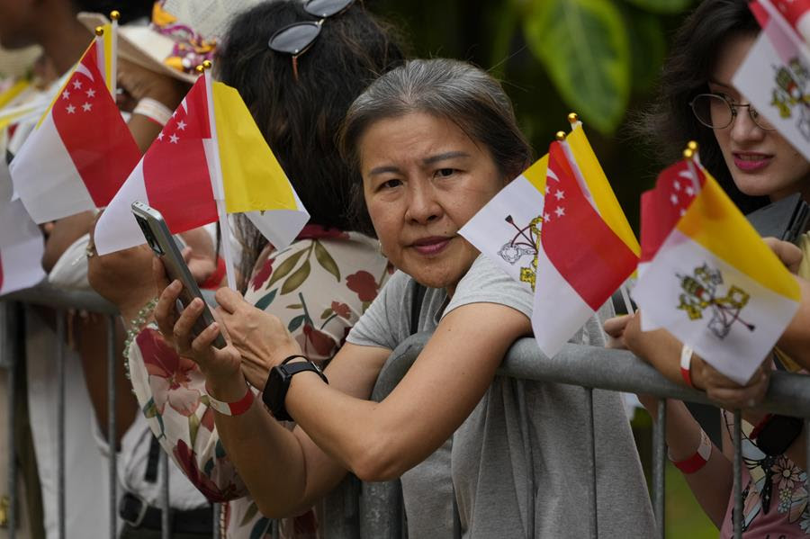 A woman leans over a metal barrier holding her phone. All around her people holding the flags of Singapore and the Vatican.