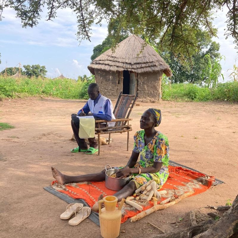 A woman sits on a mat and a man sits on a chair outside a hut.