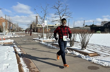 man in red and black flannel shirt, dark blue pants, black knit cap and sunglasses runs down a city sidewalk on a bright winter day