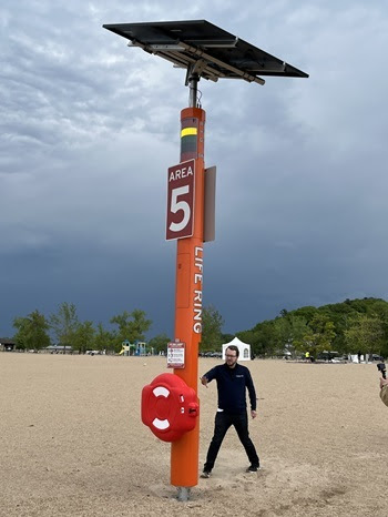 a man in black shirt and pants gestures to a tall tower on the beach. The tower is topped by red, yellow and green lights and has a life ring attached