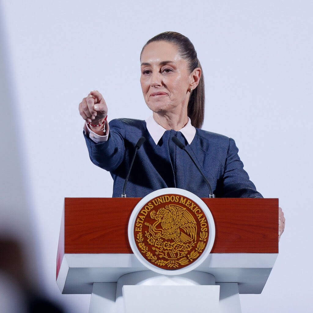 President Claudia Sheinbaum stands at a podium during a news conference and points. 