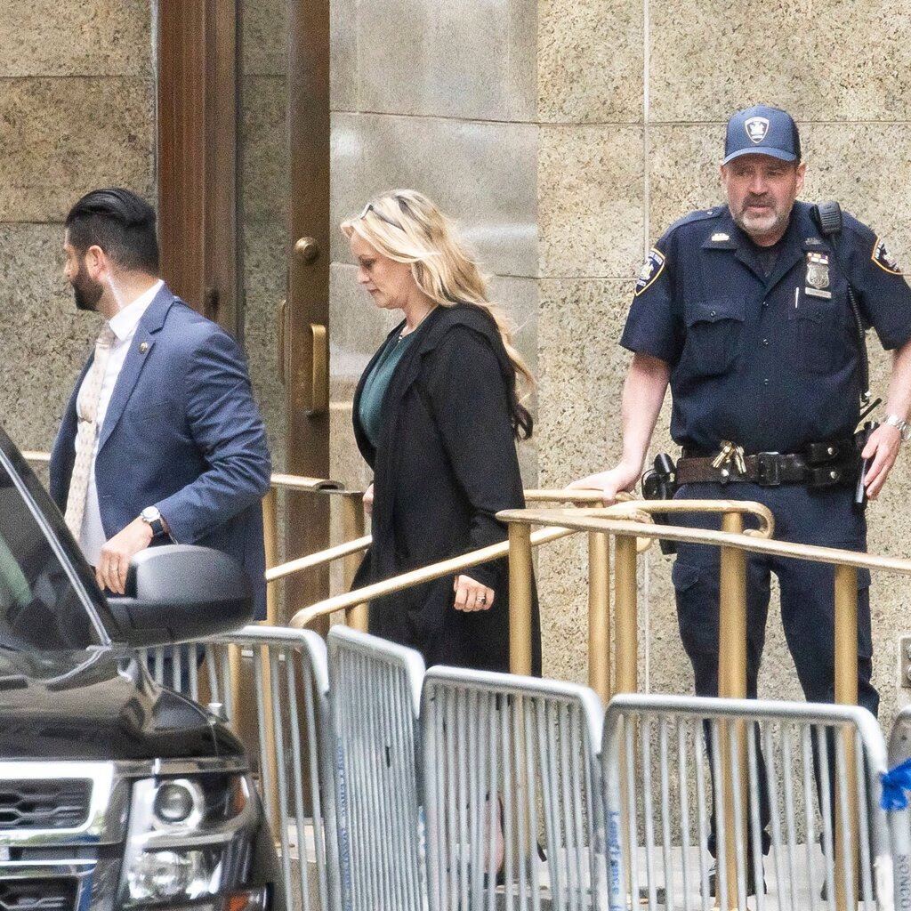 A blonde woman in a black jacket walks looking down toward a car. A police officer stands near her, and there’s a man facing away. Railing is all around.