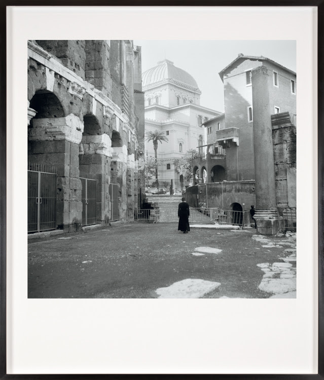 Color image of a framed black and white photograph of a woman in a black dress standing alone in a courtyard 