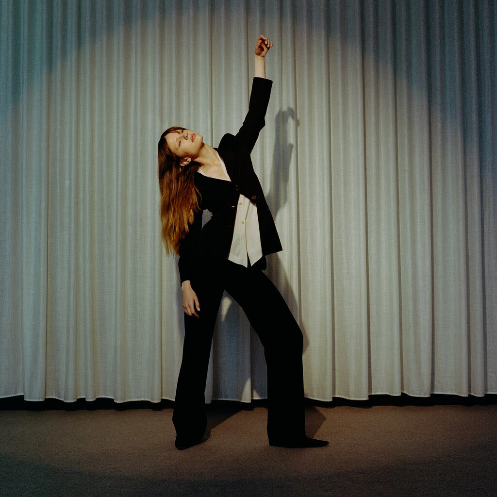 Against neutral curtains, Mia Goth strikes a post with one arm in the air, hip cocked, head titled back with long reddish-brown hair falling past the other arm.