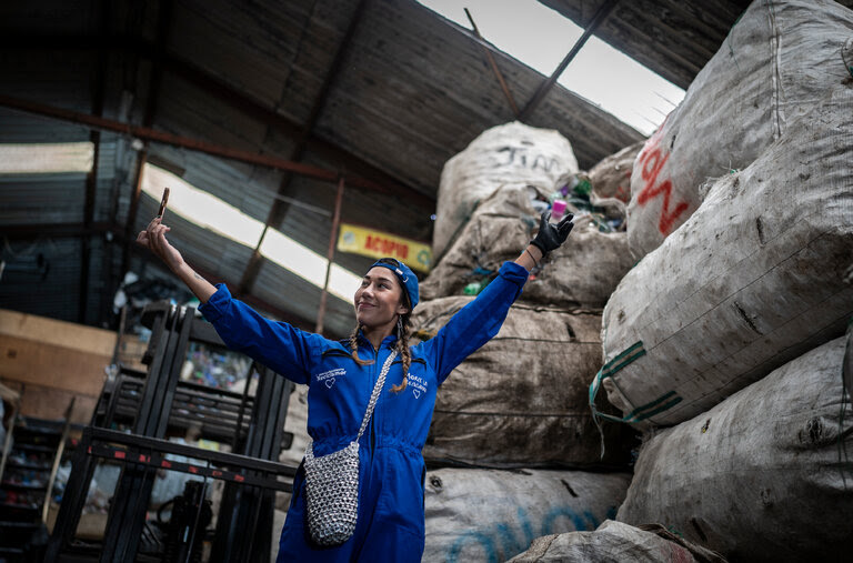 A woman in a blue jumpsuit looks at herself on a mobile phone surrounded by large white bags.