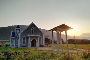 A small church building is in the foreground. The sun is seen just above the hilltops in the background.