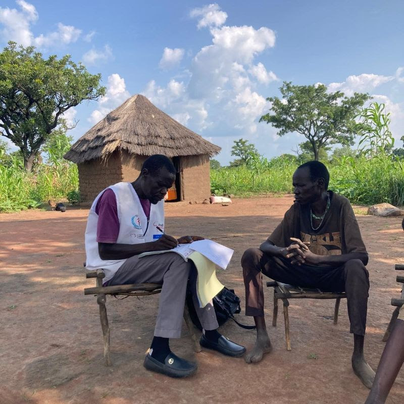 Two men seated on chairs, engaged in conversation, in front of a rustic hut surrounded by greenery.