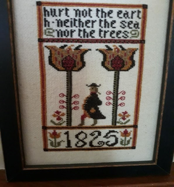 Photograph of a framed cross-stich artwork. The words at the top say "hurt not the earth - neither the sea - nor the trees" The date at the bottom is large: 1825, flanked by pink tulips. In the middle, the main image, a man in a long coat and hat stands between two large trees. 