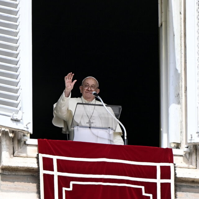 Francis, in white, gestures from an open window, with a lectern and microphone in front of him.