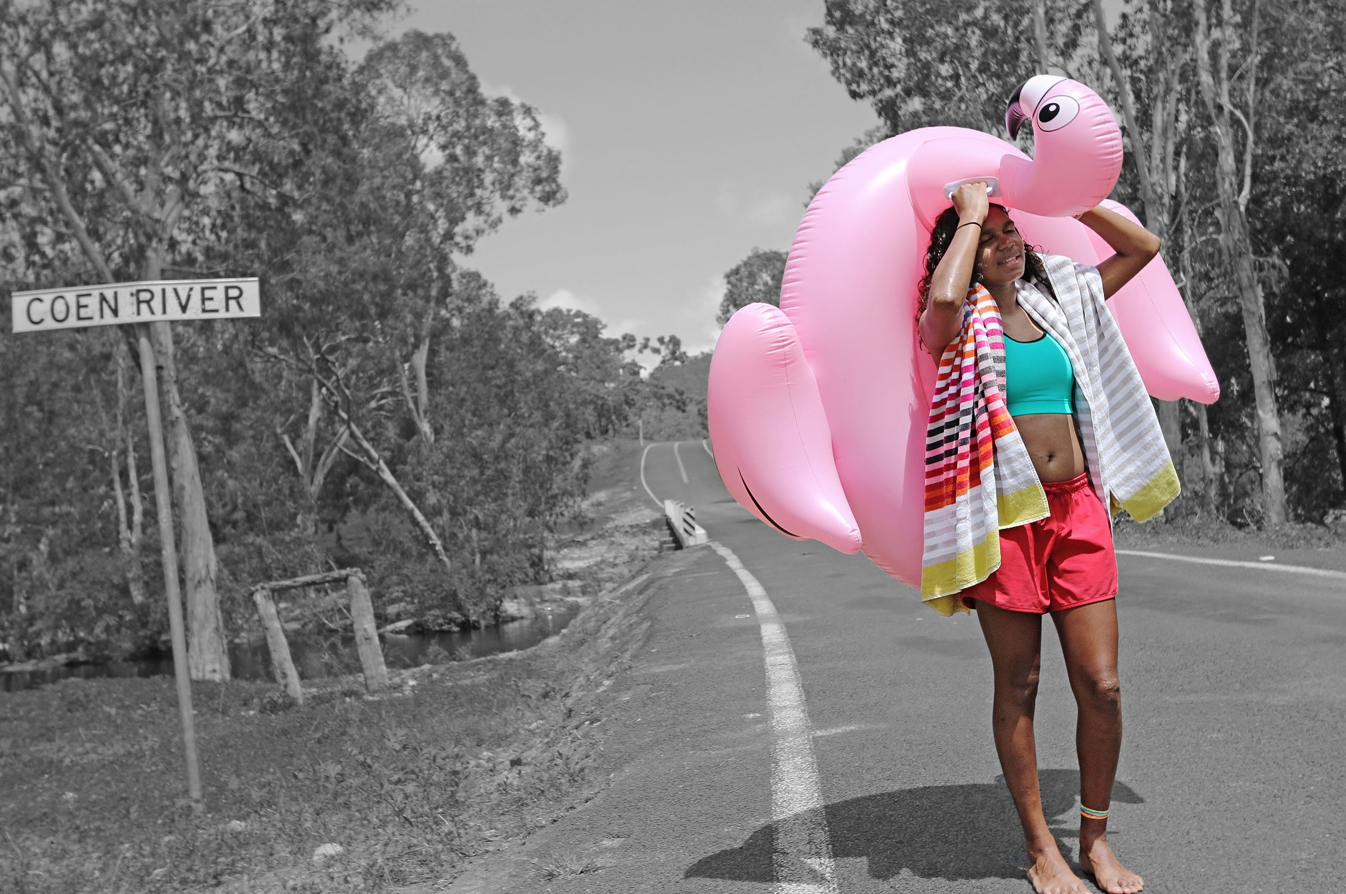 A woman carrying a pink inflatable bird behind her back and head, walking along a road with a road sign Coen River.