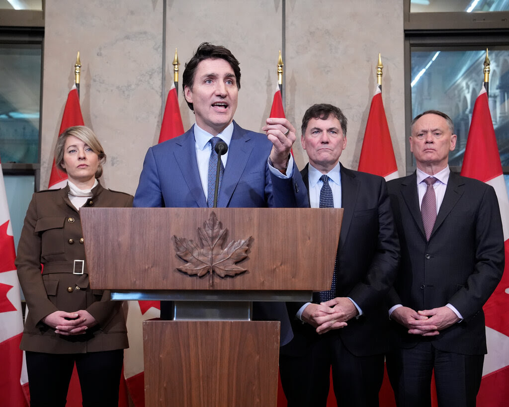 Prime Minister Justin Trudeau raises his hand while speaking at a lectern. In the background, three people stand behind him and behind them multiple Canadian flags. 