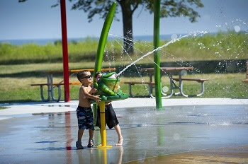 two little boys cool off and have fun playing with a spray gun on a cement water splash pad set in a grassy park area