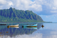 Small wooden fishing boats sit on still water near shore with a greenery covered mountain in the background. 