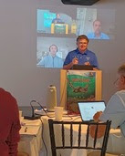 A person presents in front of a panel of people both in person and through virtual video