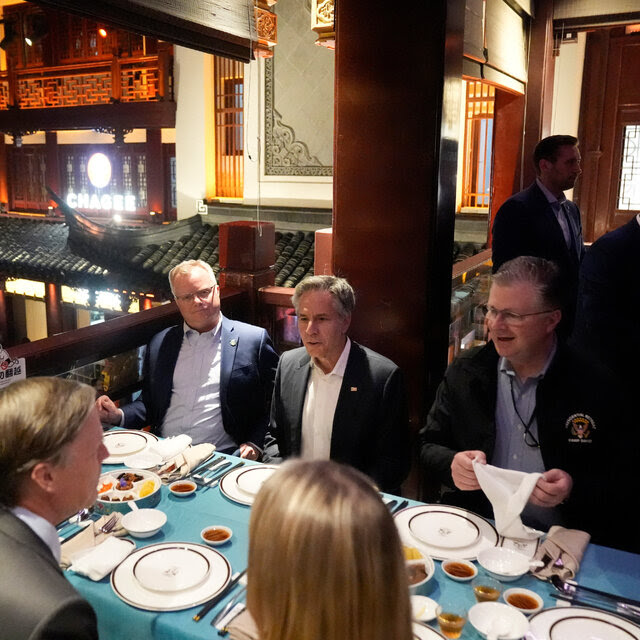 Secretary of State Antony J. Blinken, wearing a white shirt and suit jacket, sitting at a table in a restaurant with six other people dressed in formal attire. 