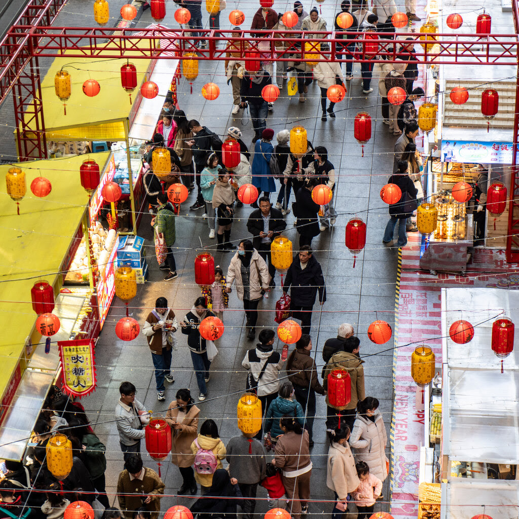 A crowd of shoppers seen from above walking between two rows of shops, one row with yellow roofs and the other with white roofs.