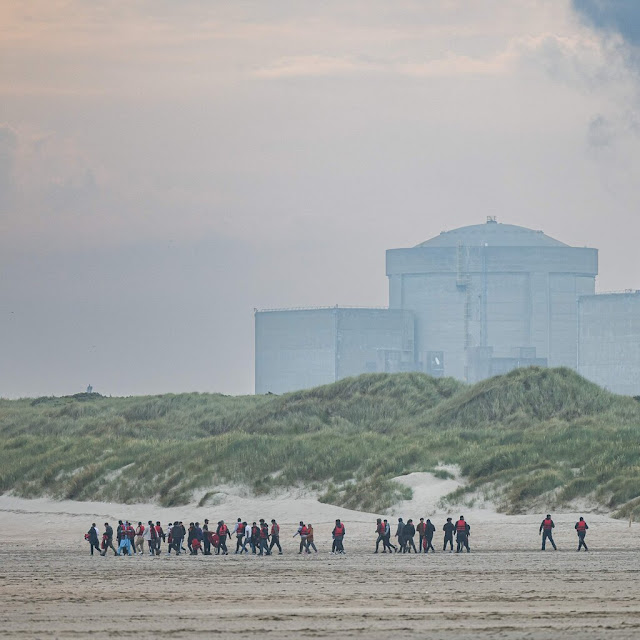 Migrants running across a beach with a nuclear power station in the background. 