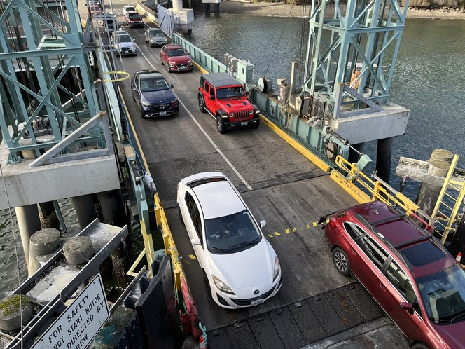 Vehicles on a bridge boarding a ferry with a dock seen in the background along with some buildings and vegetation