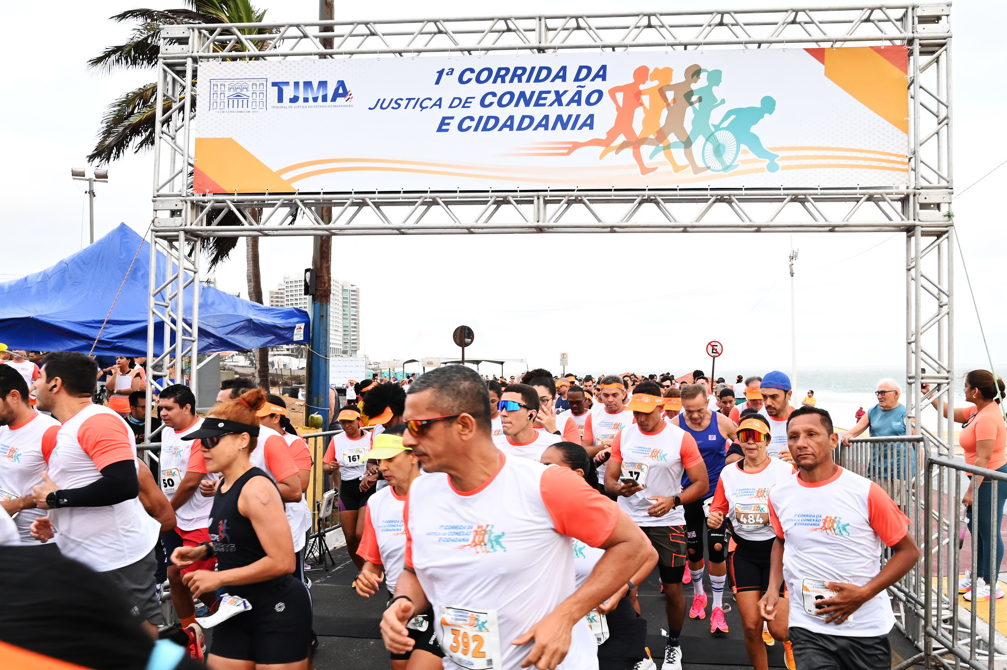 Foto colorida mostra pessoas correndo na Av Litorânea. Acima, banner da corrida do TJMA.