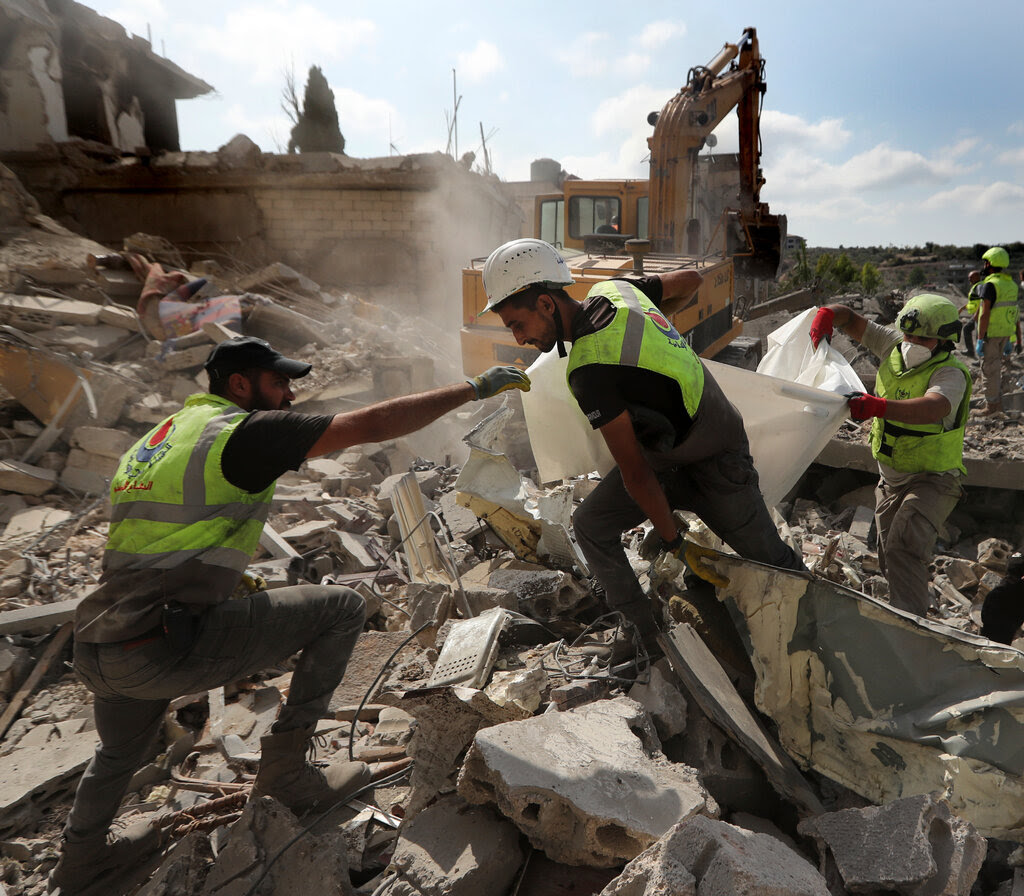 Rescue workers climb over debris while carrying a white body bag.