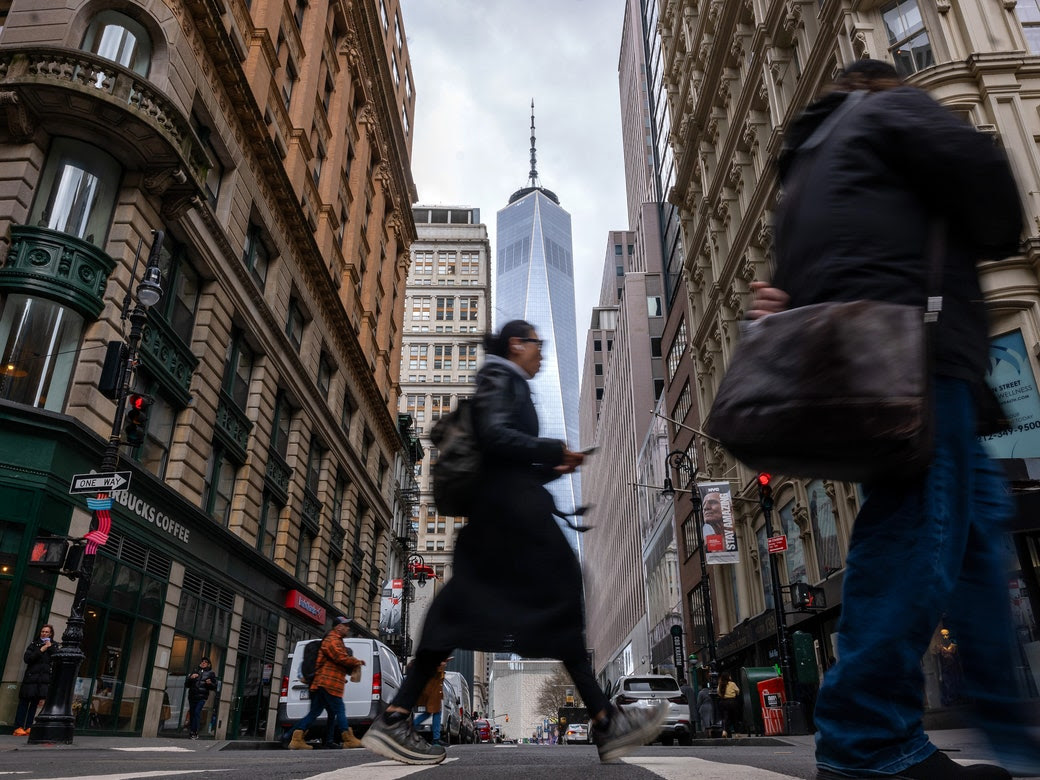 A person crosses a street in lower Manhattan with the 1 World Trade building in the background.