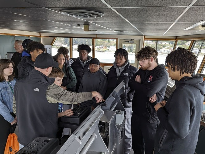 Several high schoolers listening to a crewmember speak in the wheelhouse of a ferry