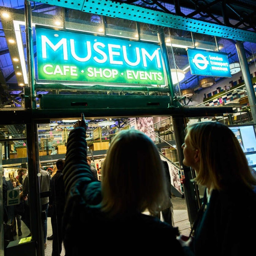 Two people at the entrance of the museum point towards the illuminated museum signage