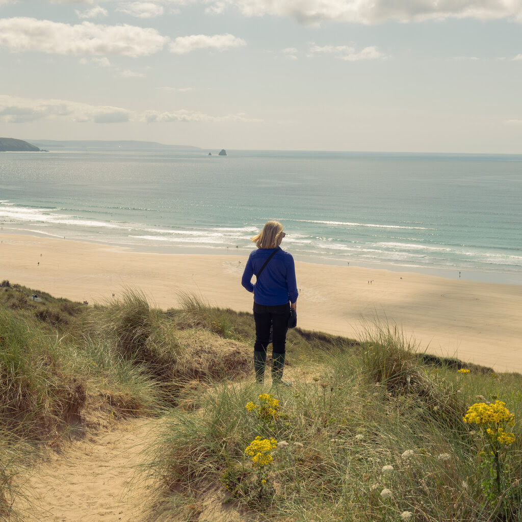 A woman turning her back to the camera on the dunes of a beach. The water is visible in the background and the dunes are grassy. There are flowers. 