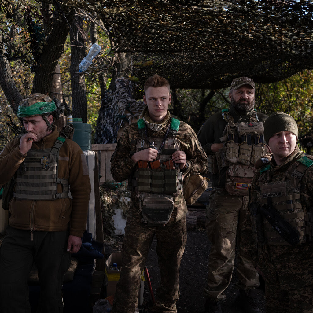 A group of soldiers in uniform stand under a camouflage canvas in the woods.