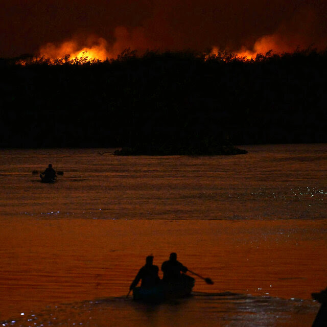 Wildfires burning in the distance, with the river in the foreground. There are two canoes of people in the river.