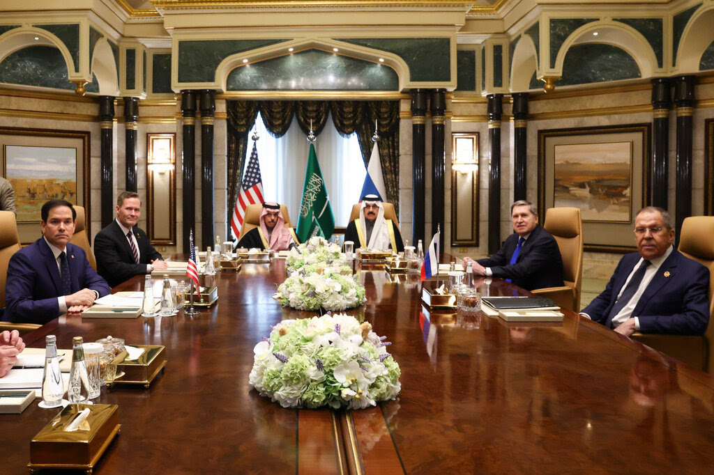 Several men sit around a table with the flags of the United States, Saudi Arabia and Russia behind them.