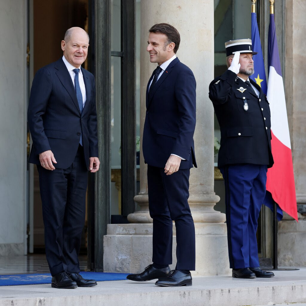 Olaf Scholz and Emmanuel Macron on the steps of the Élysée Palace, with guards in dress uniform.