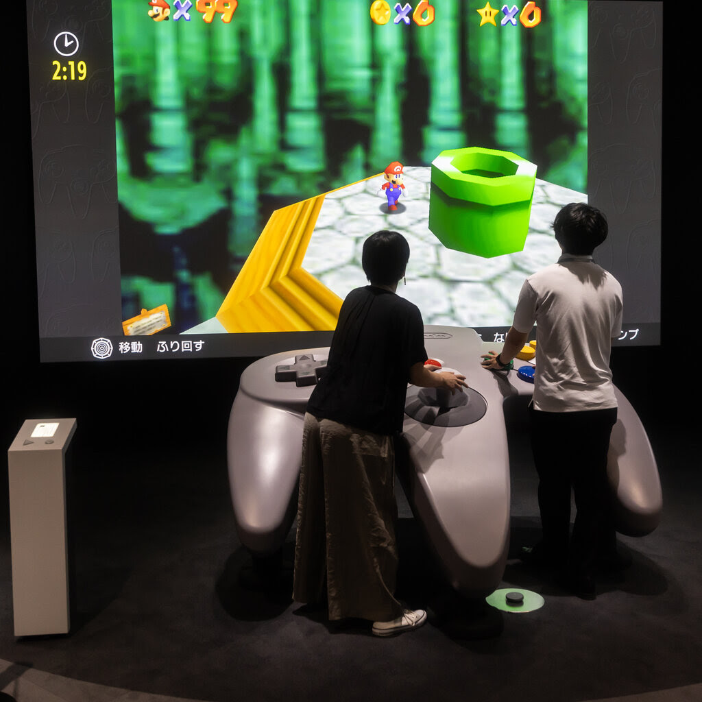 Two people operate a giant Nintendo 64 controller while playing a Mario video game.