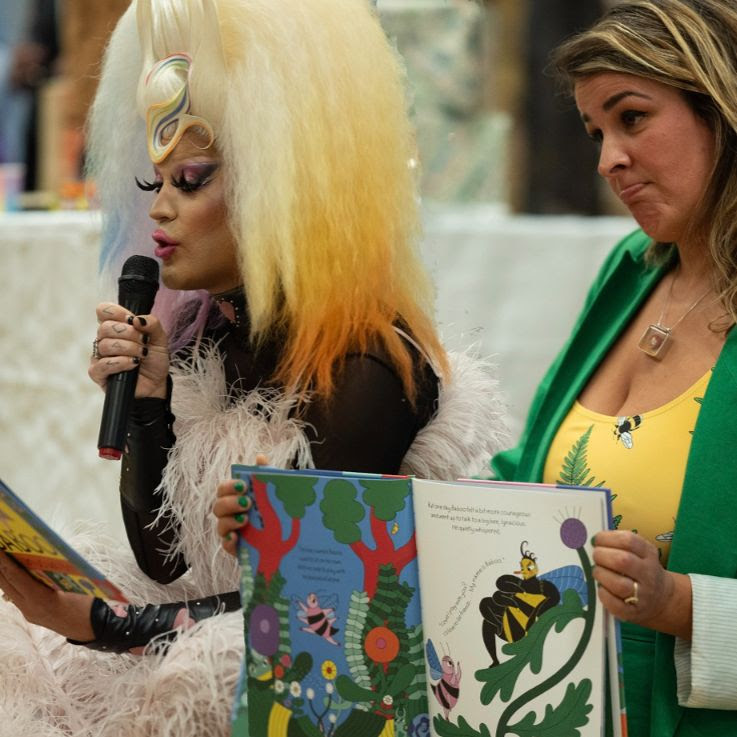 A drag queen talks into a microphone whilst a woman holds up an open book