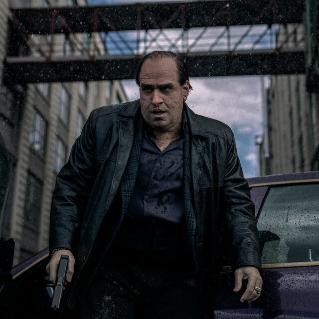 A man in a black leather jacket holding a pistol scowls in the rain