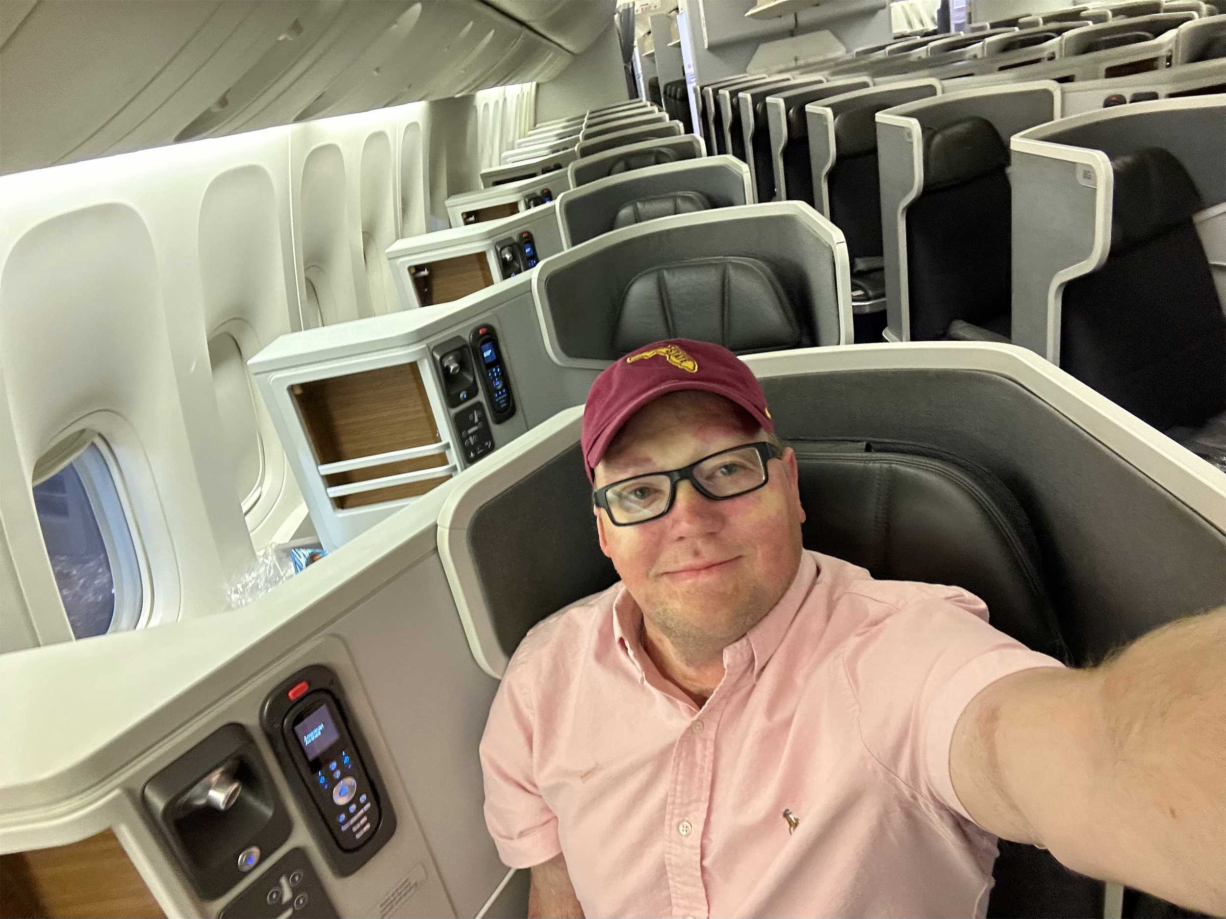 Selfie of John wearing a pink shirt and garnet baseball cap seated in business class on a Boeing 777 aircraft. Selfie of John wearing a pink shirt and garnet baseball cap seated in business class on a Boeing 777 aircraft.