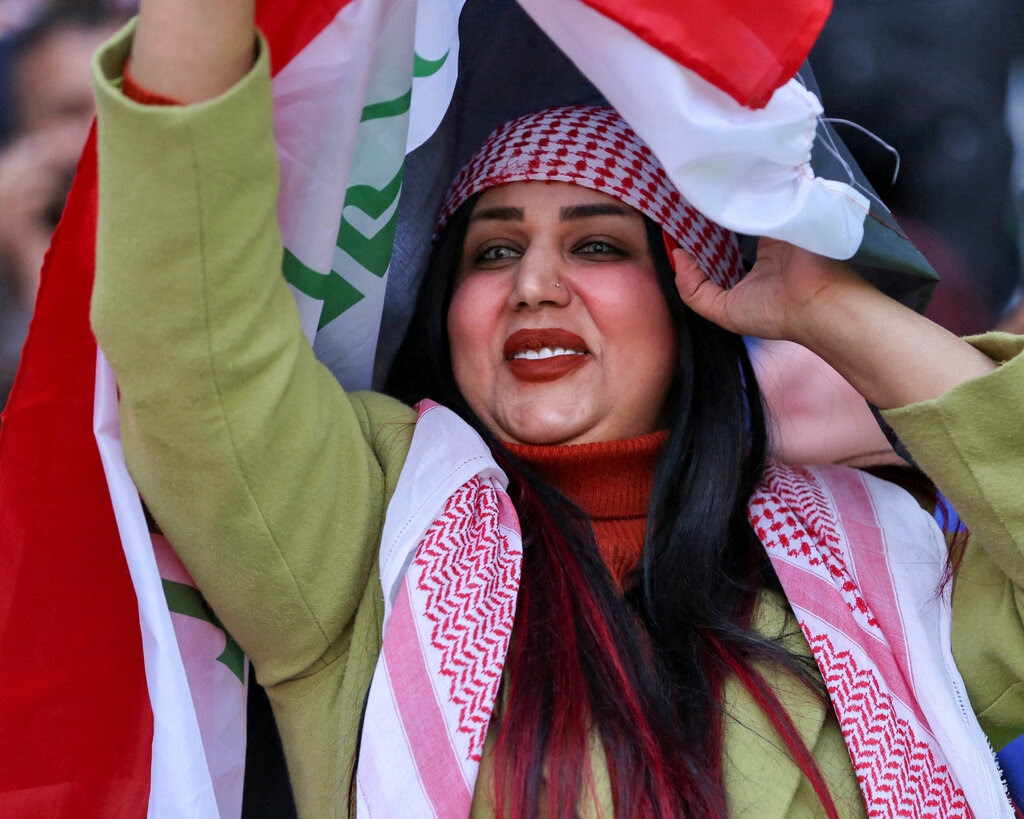 A woman wearing a green coat and red head scarf holding Iraqi flag. 