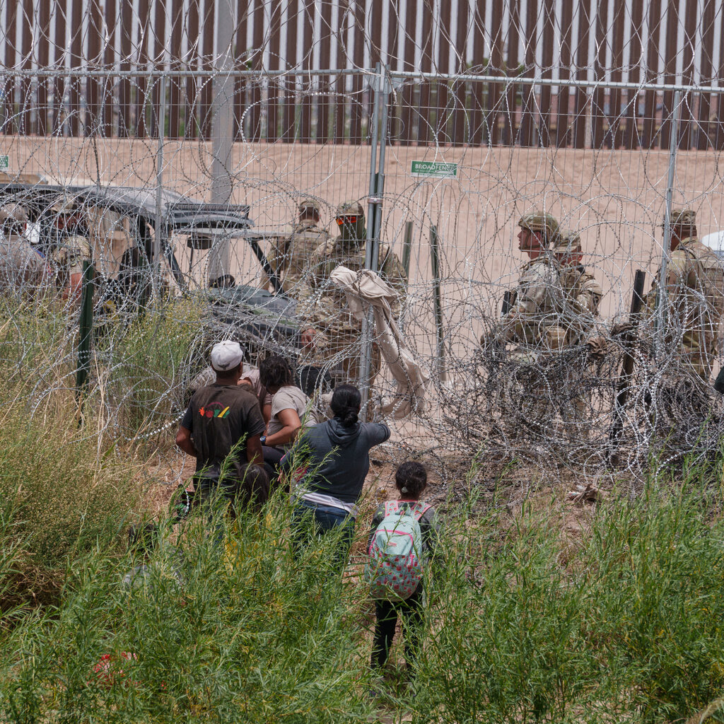 A group of migrants, including a small child with a backpack, look at border agents in fatigues across a border fence covered in concertina wire.